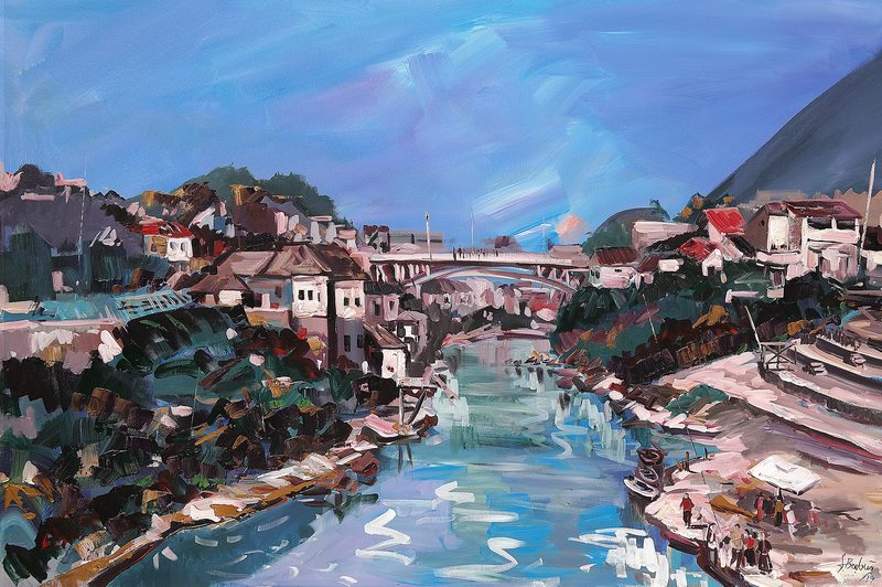 Red-roofed houses on both banks of a river, a bridge further on and a green mountainside behind; blue, pink and green tones.