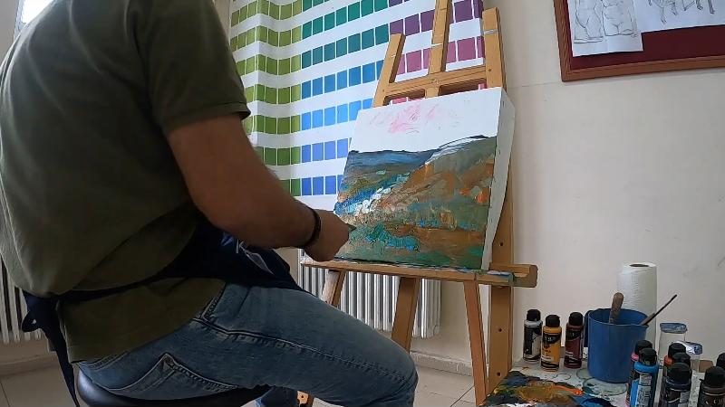 Çalıştay atölyesinde şövalenin önünde oturan ressam arkadan görünüyor; tuvalde mavi, yeşil ve okra bir manzara, masada boya şişeleri.